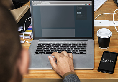 Black person typing on a laptop, at a wooden table with notebooks, coffee cup, and phone