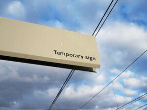 A cream colored billboard interrupts a cloudy blue sky. The billboard reads, Temporary Sign.