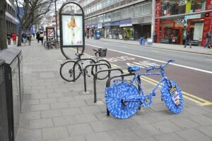 A UK city street lined with stores. On the sidewalk running down the left side of the image stands a bike stand with a bike wrapped in blue paper with the words The Electric repeated all over it. A similar bike is on the right side of the street as well.